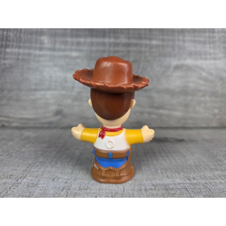May include: A small plastic toy figure of a cowboy character, wearing a brown hat, yellow shirt, blue pants, and a red neckerchief. The figure has outstretched arms and is standing on a gray surface.