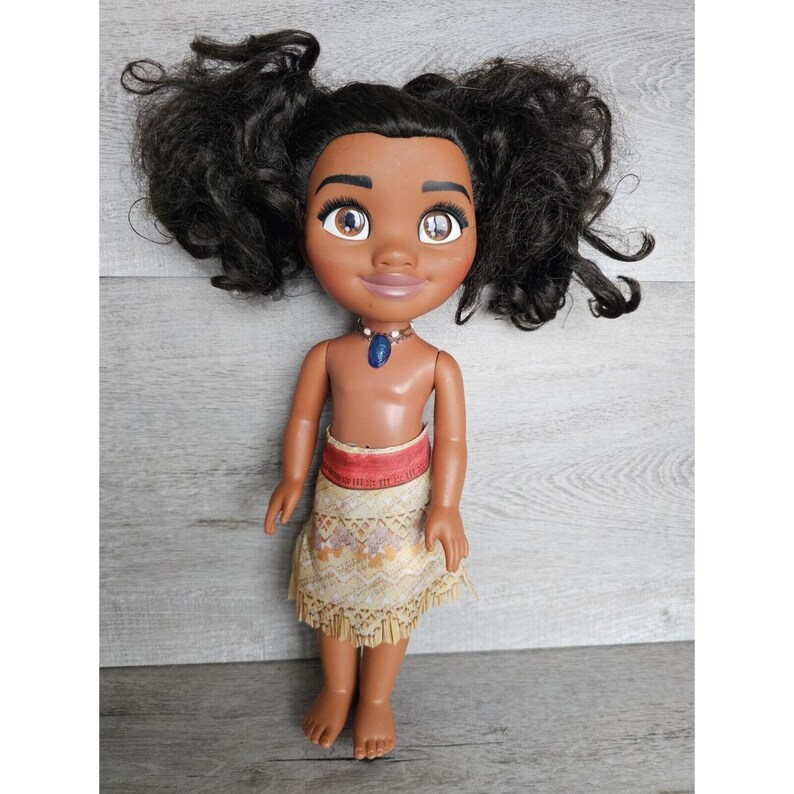 May include: A brown-skinned doll with black curly hair styled in pigtails. The doll is wearing a brown and gold patterned skirt with a red waistband and a blue necklace with a blue gem pendant.