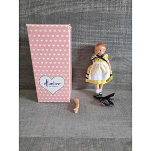May include: A vintage Effanbee doll with a yellow and white dress, black shoes, and red hair, next to its pink box with white heart patterns. The doll's arm is detached. The box has the Effanbee doll company logo.