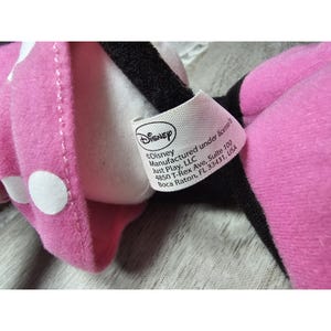 May include: Close-up of a pink plush toy with white polka dots and a black bow. A white tag with the Disney logo and text is attached. The tag reads "Disney Manufactured under license by Just Play, LLC".