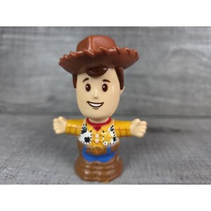 May include: A small, plastic toy figure of a cowboy character. The figure has a brown cowboy hat, a yellow shirt with black and white cow-print accents, blue pants, and a red neckerchief. The figure's arms are outstretched.
