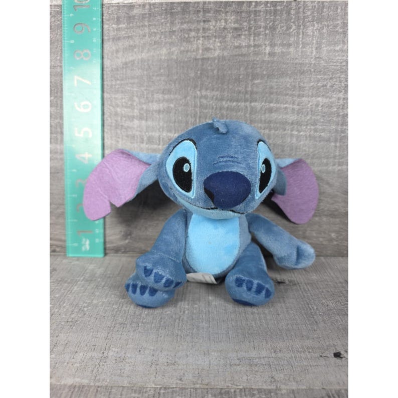 May include: A blue plush toy character with large ears, a light blue chest patch, and dark blue nose, positioned next to a green ruler showing a height of approximately 6 inches. The character has embroidered black eyes and blue paw details.