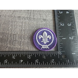 May include: Purple embroidered scout patch with a white fleur-de-lis and a white rope design. The patch is approximately 1 inch in diameter.