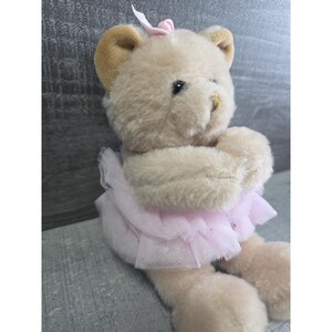 Pu&ograve; includere: Un orsacchiotto di peluche in un costume da ballerina. L'orso &egrave; marrone chiaro con orecchie marroni e un fiocco rosa. Indossa un tut&ugrave; di tulle rosa. Le braccia dell'orso sono incrociate davanti al petto.