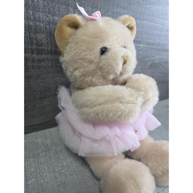 Pu&ograve; includere: Un orsacchiotto di peluche in un costume da ballerina. L'orso &egrave; marrone chiaro con orecchie marroni e un fiocco rosa. Indossa un tut&ugrave; di tulle rosa. Le braccia dell'orso sono incrociate davanti al petto.