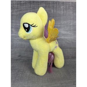 May include: A soft, yellow plush pony toy with blue eyes, black eyelashes, and a purple mane and tail. It features yellow wings and purple cross-shaped markings on its legs. The toy is designed to resemble a cartoon character.