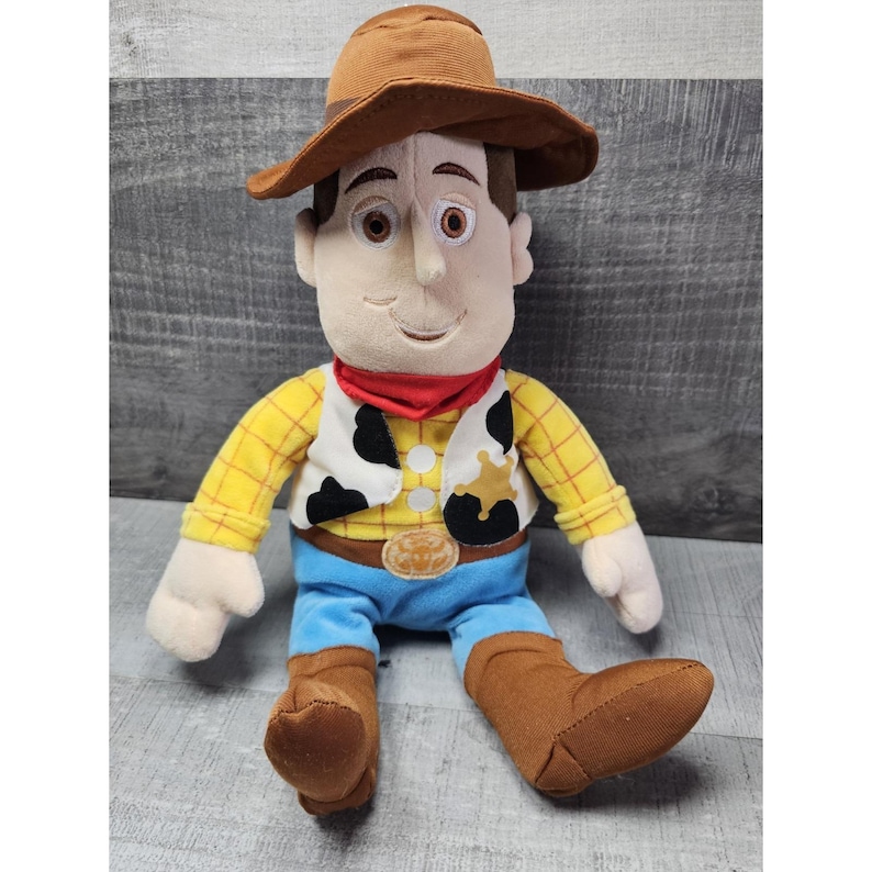May include: A plush toy of a cowboy character, wearing a brown hat, yellow plaid shirt, blue pants, and brown boots. The toy has a red bandana and a black and white vest. The character has a friendly expression.