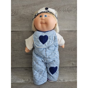 May include: A Cabbage Patch Kid doll wearing a blue and white gingham overall with blue heart accents, a white and black striped headband, and a white hat with a blue button.