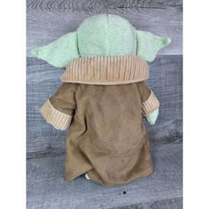 May include: A plush doll of a character with large ears and a tan coat. The doll has a light green head and ears, a tan collar, and a brown coat. The doll is made of soft, plush material.