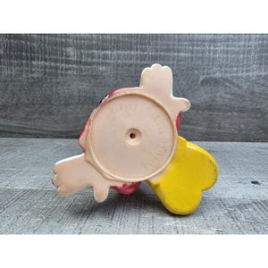 May include: A vintage, bottom-view of a Disney character figurine. The figurine is primarily pink, red, and yellow, with white accents. The base is circular with the text "DISNEY" and "MADE IN CHINA".