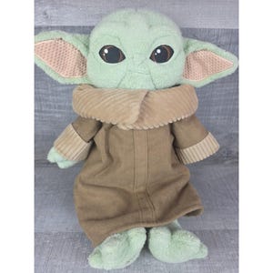 May include: A plush doll of a character with large brown eyes, large ears, and green skin. The doll wears a brown coat with a corduroy collar and has green feet and hands. The doll is a popular character from a science fiction franchise.