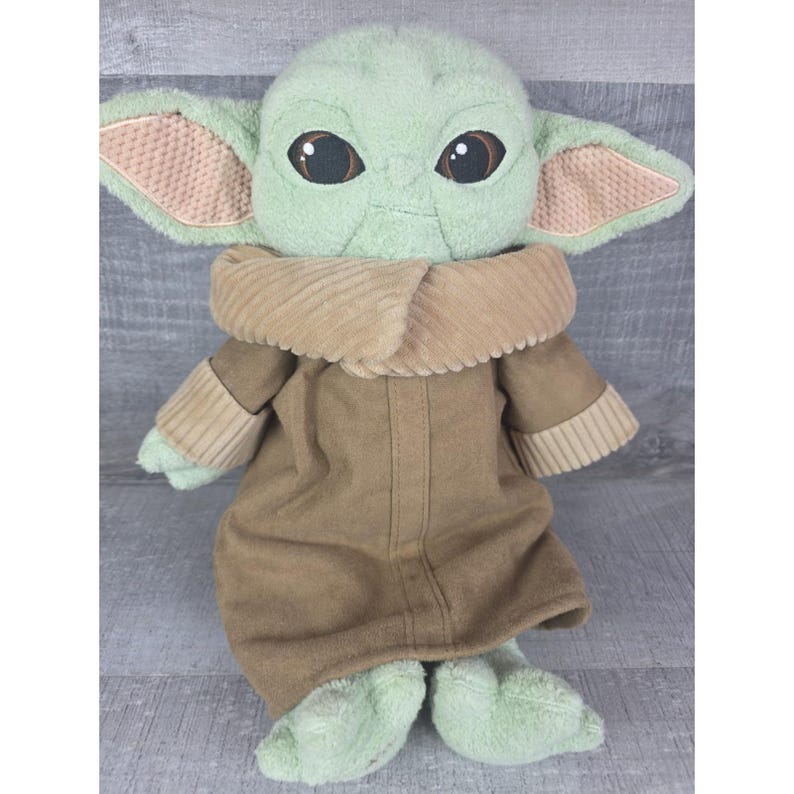 May include: A plush doll of a character with large brown eyes, large ears, and green skin. The doll wears a brown coat with a corduroy collar and has green feet and hands. The doll is a popular character from a science fiction franchise.