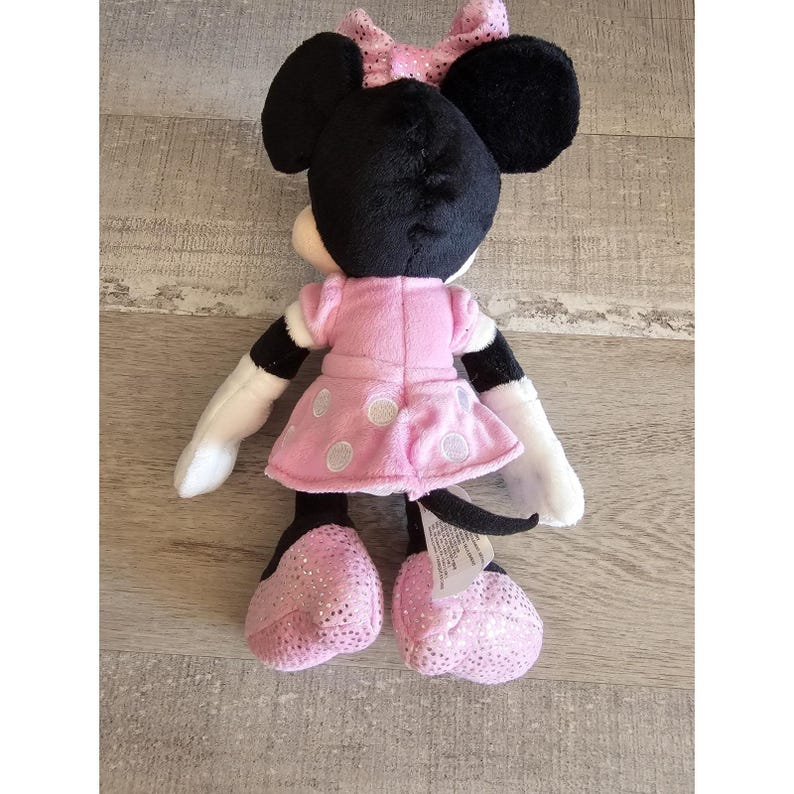 May include: A plush toy of Minnie Mouse wearing a pink polka dot dress with a black bow on her head. She has black ears and a black nose.