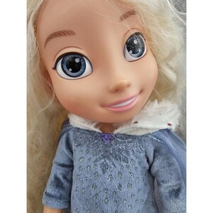 May include: A blonde doll with blue eyes and a light blue dress with a snowflake pattern. The doll is wearing a white fur-trimmed hood.