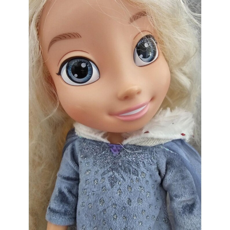 May include: A blonde doll with blue eyes and a light blue dress with a snowflake pattern. The doll is wearing a white fur-trimmed hood.