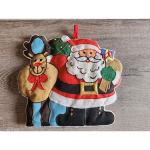 May include: A red and white Santa Claus ornament with a brown reindeer holding a green and red gift bag. The ornament is made of fabric and has embroidered details.