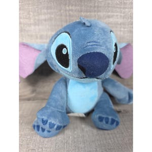 May include: A plush toy of the character Stitch, featuring soft blue fur, large pointed ears with light purple inner sections, a dark blue nose, and embroidered black eyes with white highlights. The character has a light blue belly patch.