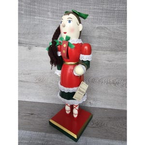 May include: A wooden nutcracker figurine of a girl wearing a red dress with white lace trim, a green sash, and a green bow in her hair. She is holding a white ball in her right hand. The nutcracker is standing on a red and green base with a gold trim. The base has a label that reads "Original German Nutcracker Collection".