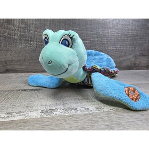 Op de afbeelding: Een pluche schildpad in blauwe en groene tinten. De schildpad heeft een lachend gezicht, grote ogen met wimpers en een patroon op de schild. De vinnen hebben een bruin en beige ontwerp. Het speelgoed is gemaakt van zacht, pluche materiaal.