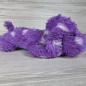 以下が含まれることがあります： 白い斑点のある紫色のシャギーファーで覆われた、犬の形をしたぬいぐるみ。このおもちゃには、小さな直立した尻尾、垂れ耳、滑らかな薄紫色の鼻が特徴です。2つの小さな丸い黒いプラスチック製の目が付いています。