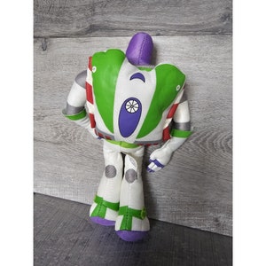 Peut inclure: Un jouet en peluche de Buzz l'&Eacute;clair de la franchise Toy Story. Le jouet est vert, blanc et violet avec une bande rouge sur le bras. Le jouet est assis sur une surface en bois.