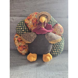 May include: A plush turkey with a brown and white checkered body, a red and white polka dot scarf, and a straw hat. The turkey's tail feathers are made of fabric with a pumpkin and plaid pattern.