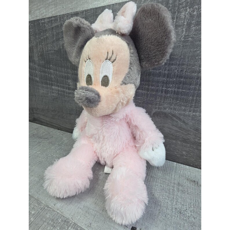 Puede incluir: Un peluche de Minnie Mouse sentada. La mu&ntilde;eca tiene un cuerpo rosa, orejas grises y un lazo rosa. La cara es rosa claro con ojos bordados y una nariz gris. La mu&ntilde;eca est&aacute; hecha de un material suave y difuso.