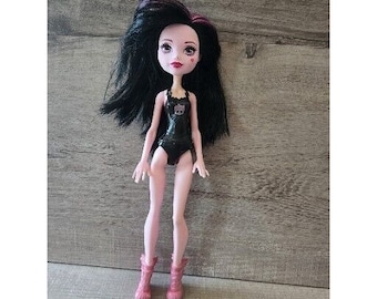 May include: A doll with long black hair with purple streaks, wearing a black swimsuit and pink boots. The doll has pale skin, red lips, and a small heart-shaped mark on its cheek. The swimsuit features a skull design.