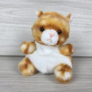 May include: A plush toy shaped like a small cat with orange and white fur. The cat has a white muzzle, a small pink nose, and dark eyes. Its body is primarily orange with a large white patch covering the chest and belly area, and it has small ears.