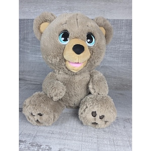 May include: A plush teddy bear with a light brown fur. The bear has large blue eyes, a tan snout, and a pink mouth. The bear is sitting upright with its arms and legs extended.