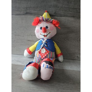 Könnte beinhalten: Eine Plüsch-Clown-Puppe mit einem roten, gelben und blauen Outfit. Der Clown hat eine große rote Nase, einen weißen Knopf auf seiner Weste und ein gestreiftes Muster auf seiner Hose. Die Schuhe des Clowns haben die Zahlen 1, 2, 3 und 4 darauf.