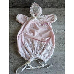 May include: A vintage baby romper in pale pink with a matching bonnet. The romper has short sleeves, a lace-trimmed hem, and a small embroidered detail. The bonnet is white with a floral pattern and a delicate bow.