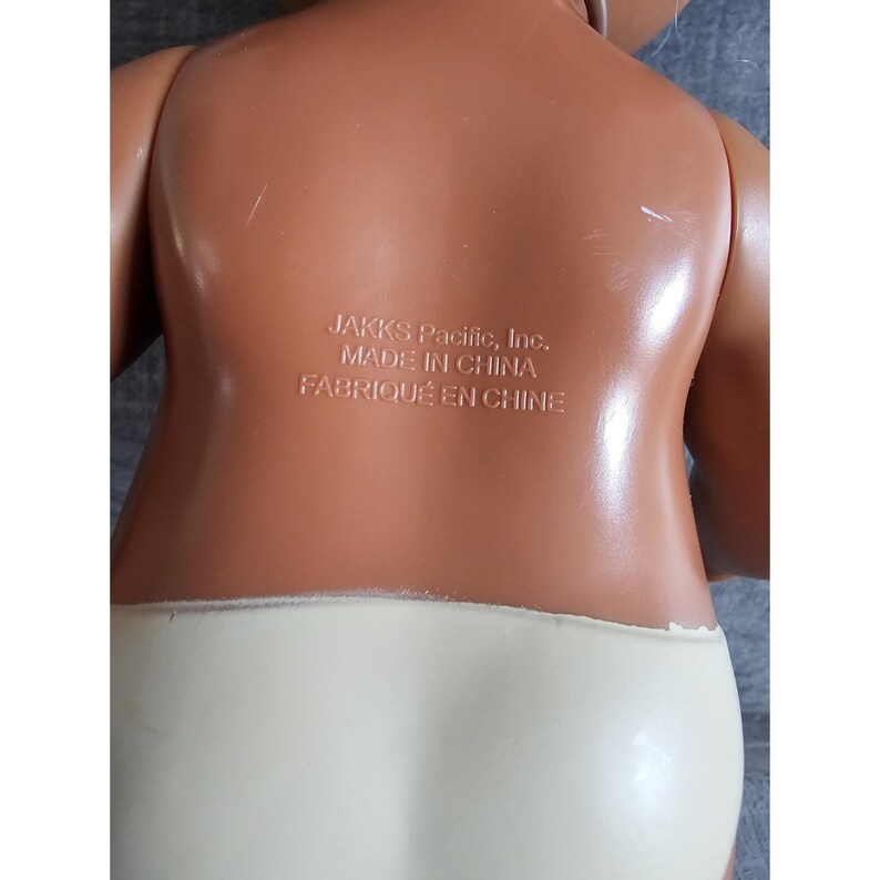 Pu&ograve; includere: Una bambola di plastica marrone con un pannolino bianco. La bambola ha un'etichetta sul retro che recita "JAKKS Pacific, Inc. MADE IN CHINA FABRIQU&Eacute; EN CHINE".