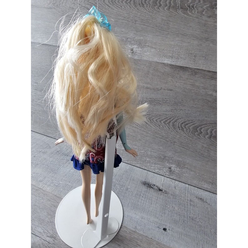 Puede incluir: Una mu&ntilde;eca rubia con un lazo azul en el pelo, vestida con un vestido estampado en rojo, azul y blanco, de pie sobre un soporte de mu&ntilde;eca blanco.