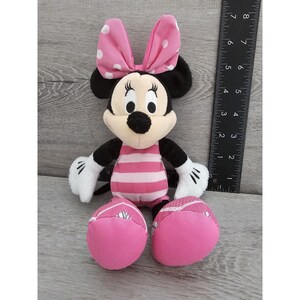 May include: A pink and black plush toy of Minnie Mouse wearing a pink and white polka dot bow and a pink and white striped swimsuit.