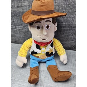 May include: A plush toy of a cowboy character, featuring a brown hat, a yellow plaid shirt, a white vest with black spots, blue pants, and brown boots. The toy has a red neckerchief and a sheriff's badge.