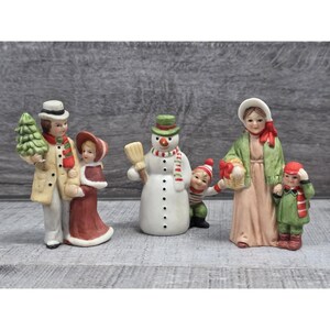 May include: A collection of ceramic Christmas figurines. The figurines include a family, a snowman with a broom, and a woman with two children. The family is dressed in winter attire, and the snowman has a scarf. The figurines are painted in various colors.