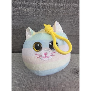 Puede incluir: Un llavero de felpa con forma de gato en colores pastel. El gato tiene ojos grandes y brillantes, una nariz y boca bordadas en rosa y un clip de plástico amarillo. Las orejas del gato tienen detalles brillantes. El llavero está diseñado para sujetarse a una mochila o bolso.