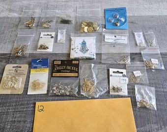 May include: Assortment of small plastic bags filled with various jewelry making supplies, including earring hooks, jump rings, beads, and findings. The bags are arranged on a brown surface with a brown envelope labeled "12".