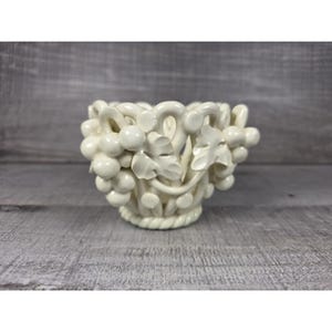May include: A cream-colored ceramic bowl featuring an intricate openwork design of sculpted grape clusters and leaves. The base of the bowl is finished with a decorative rope-style border, and the entire piece has a smooth, glossy finish.