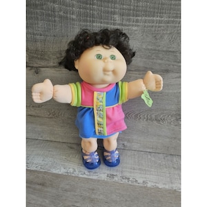 May include: A Cabbage Patch Kid doll with brown curly hair, green eyes, and a pink, blue, and green dress. The doll is wearing blue jelly shoes and has a tag attached to its wrist.