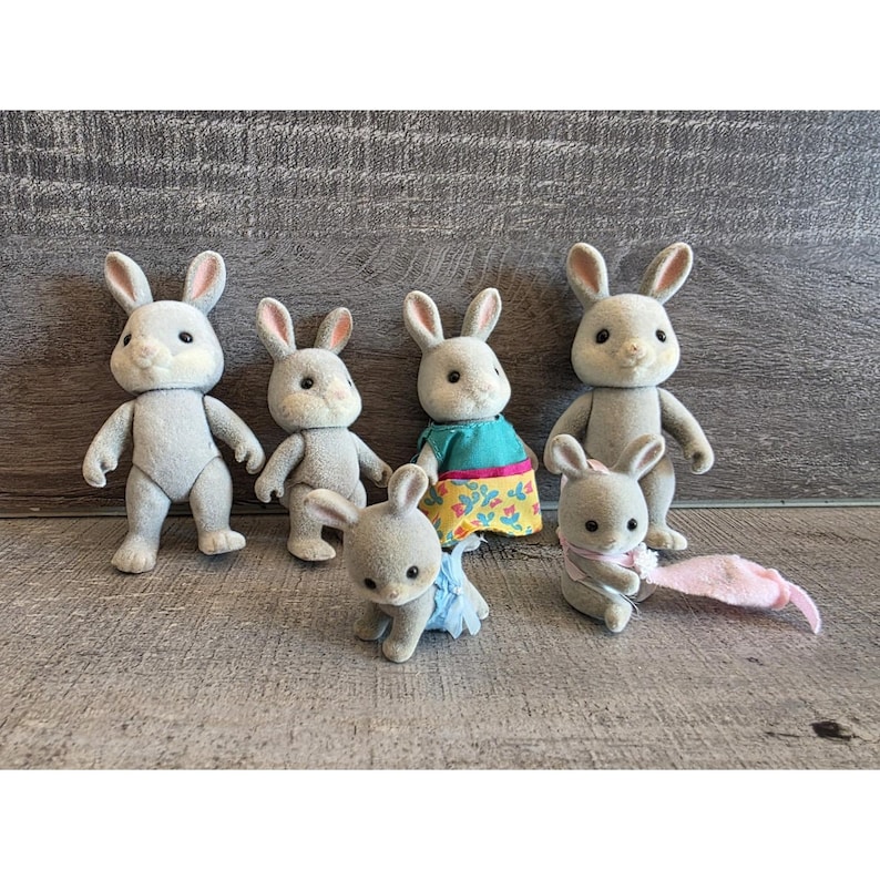 Puede incluir: Un conjunto de seis figuras de conejos grises. Algunos conejos est&aacute;n de pie, otros sentados. Uno de ellos lleva un vestido azul y amarillo. Uno tiene una bufanda rosa. Sobre una superficie de madera.