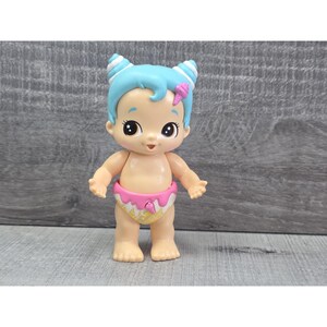 May include: A small, light-skinned doll with blue hair styled in two buns with white stripes. The doll has large eyes and a pink bow. It wears a pink and yellow diaper with an ice cream cone design. The doll is standing.