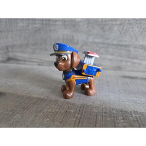 Puede incluir: Un perro de juguete de plástico marrón que lleva un gorro y uniforme de policía azules. El perro tiene una mochila con un elemento rojo y blanco. El juguete está sobre una superficie gris.