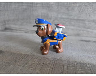 May include: A brown plastic toy dog wearing a blue police hat and uniform. The dog has a backpack with a red and white element. The toy is standing on a gray surface.