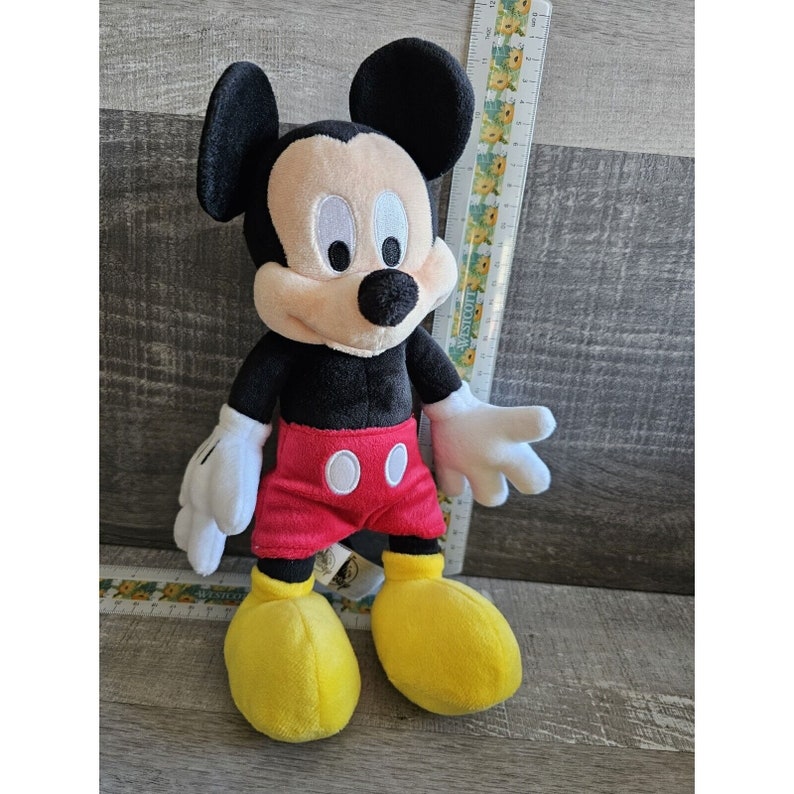 Puede incluir: Un juguete de peluche Mickey Mouse negro con pantalones cortos rojos y zapatos amarillos. El juguete est&aacute; de pie sobre una superficie de madera.