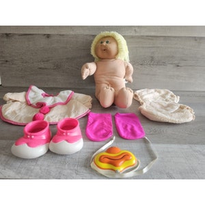 以下が含まれることがあります： ブロンドの髪とピンクと白の衣装を着たキャベツパッチキッズ人形。人形はピンクの靴、ピンクのレッグウォーマー、白いおむつを履いています。人形は黄色とオレンジの哺乳瓶も持っています。