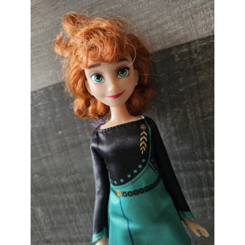 May include: A red-haired doll wearing a black and green dress with gold accents. The doll has freckles and green eyes.