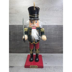Puede incluir: Figura de soldado cascanueces de madera, de pie sobre una base roja. La figura lleva un sombrero negro, una chaqueta verde con ribetes dorados, pantalones rojos y botas negras. Sostiene una espada plateada y tiene barba y bigote blancos. La base dice "2003".