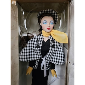 Könnte beinhalten: Eine Porzellanpuppe, die einen schwarz-weiß karierten Umhang, ein schwarzes Kleid, gelbe Handschuhe und einen gelben Schal trägt. Die Puppe hat braunes Haar, blaue Augen und roten Lippenstift.